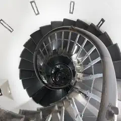 A top-down view of a black spiral staircase with a metal handrail, set within a white-walled space.
