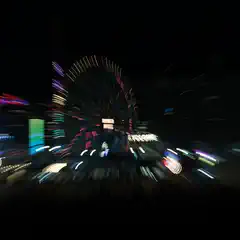 A nighttime zoom burst captures a brightly lit Ferris wheel and surrounding amusement park lights as streaks radiating from the center.