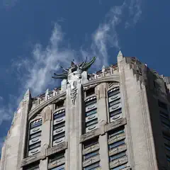 A tall Art Deco building with a prominent winged sculpture at its crown rises against a blue sky with scattered clouds.