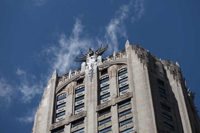 A tall Art Deco building with a prominent winged sculpture at its crown rises against a blue sky with scattered clouds.