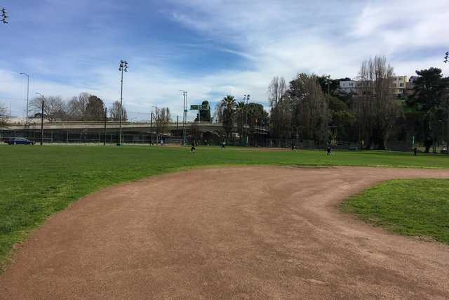 A baseball diamond occupies a grassy field with people scattered around it, and a bridge is visible in the background.