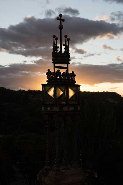 A tower with intricate designs and a cross on top, silhouetted against a cloudy sky at sunset.