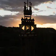 A tower with intricate designs and a cross on top, silhouetted against a cloudy sky at sunset.
