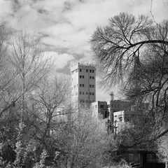 A large industrial building with multiple levels and a prominent tower, surrounded by bare trees and partially obscured by branches.