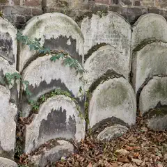 Weathered gravestones with faded inscriptions are arranged in rows against a stone wall, partially covered by moss and ivy.
