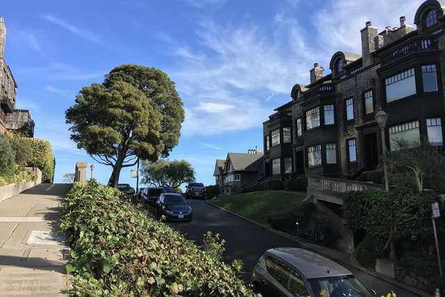 An urban area featuring steeply sloping streets lined with residential homes and tall trees, set against a backdrop of a bright blue sky with wispy clouds.