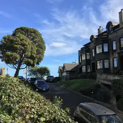 An urban area featuring steeply sloping streets lined with residential homes and tall trees, set against a backdrop of a bright blue sky with wispy clouds.