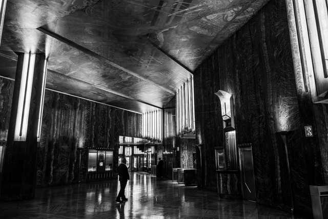 A lone figure walks through a grand, dimly lit lobby with polished marble walls, illuminated columns, and a detailed ceiling mural.