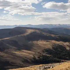 A mountain range stretches out under a blue sky dotted with clouds, featuring grassy hills and trees in varying shades of green and brown.