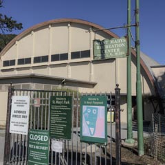 A cream-colored building with a curved roof. A green sign on a pole reads "Saint Mary's Recreation Center." A metal gate blocks the entrance with multiple signs attached.