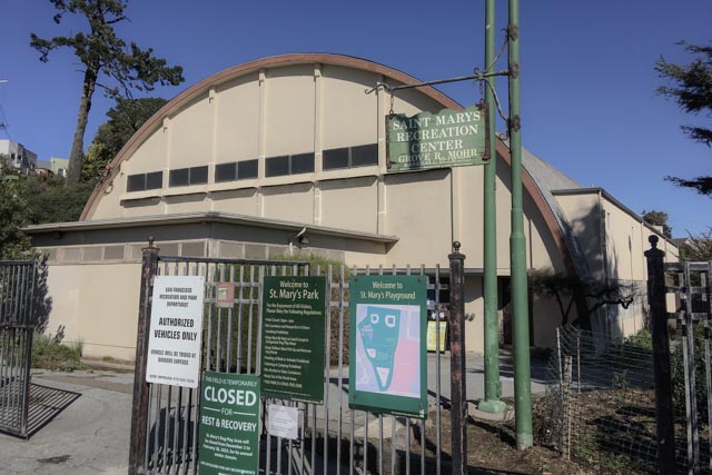 A cream-colored building with a curved roof. A green sign on a pole reads "Saint Mary's Recreation Center." A metal gate blocks the entrance with multiple signs attached.