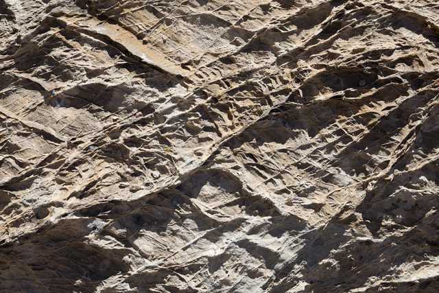 A rocky cliff face, showing layers and crevices, with sunlight casting shadows on its surface.