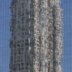 A tall glass building reflects the distorted image of a neighboring structure against a clear blue sky.