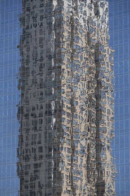 A tall glass building reflects the distorted image of a neighboring structure against a clear blue sky.