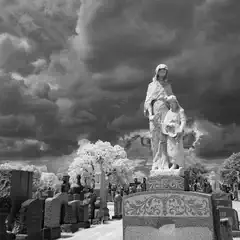An eroded statue of two figures stands prominently in a cemetery under a dramatic, cloud-filled sky.