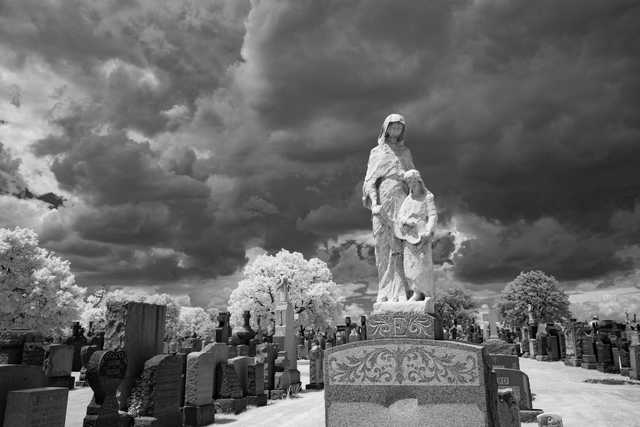 An eroded statue of two figures stands prominently in a cemetery under a dramatic, cloud-filled sky.