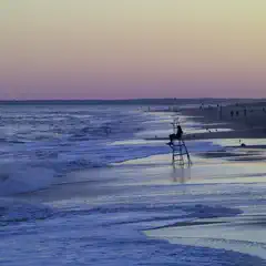 A lifeguard sits atop a tower on a beach at dusk, with waves rolling in and people scattered along the shoreline under a gradient sky.