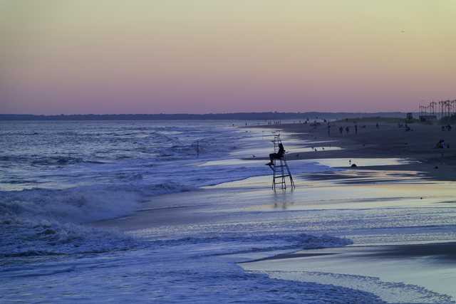 A lifeguard sits atop a tower on a beach at dusk, with waves rolling in and people scattered along the shoreline under a gradient sky.