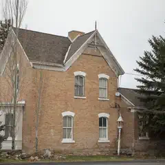 An old two-story house made of tan bricks is visible through a row of evergreen trees.