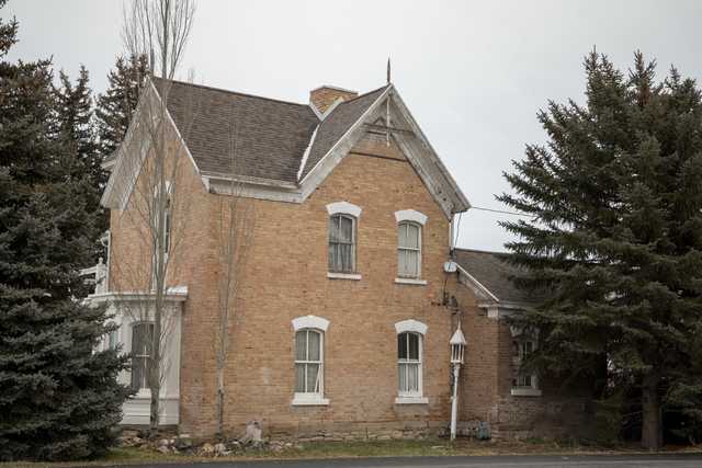 An old two-story house made of tan bricks is visible through a row of evergreen trees.