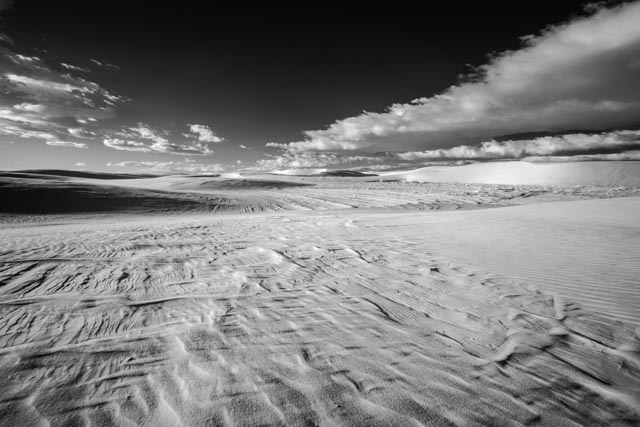 A wide expanse of rippled sand dunes stretches under a dramatic sky with scattered clouds.