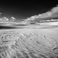 A wide expanse of rippled sand dunes stretches under a dramatic sky with scattered clouds.
