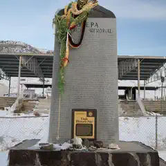 A tall stone monument with a plaque at its base and colorful leis draped around it, set against a backdrop of snow-covered mountains.