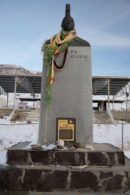A tall stone monument with a plaque at its base and colorful leis draped around it, set against a backdrop of snow-covered mountains.