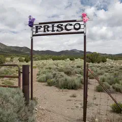 A rusted metal gate with the word "Frisco" on it. A dirt path leads through the gate into a dry, shrubby landscape with mountains in the background. Two small bouquets of flowers are attached to the top of the gate.
