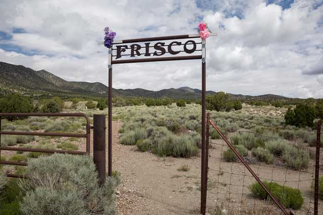 A rusted metal gate with the word "Frisco" on it. A dirt path leads through the gate into a dry, shrubby landscape with mountains in the background. Two small bouquets of flowers are attached to the top of the gate.