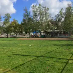 A grassy field enclosed by a fence, with trees and buildings in the background under a partly cloudy sky.