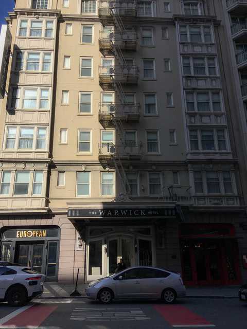 A beige hotel building with fire escapes on the facade, half in the light and half in shadow.