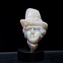 A carved opal bust of a man wearing a top hat, displayed on a black stand. The opal exhibits a subtle play of colors, giving it a luminous quality.