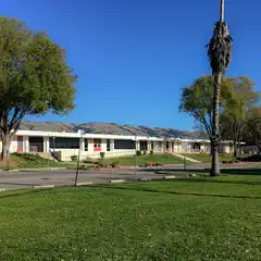 A large, low-slung building with many windows and red accents, set against a backdrop of green trees under a clear blue sky.