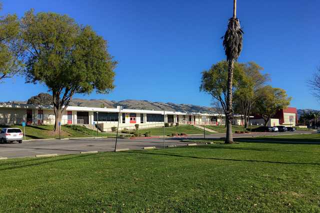 A large, low-slung building with many windows and red accents, set against a backdrop of green trees under a clear blue sky.