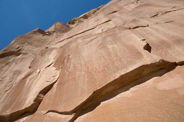 A close-up view of a rocky cliff face with visible cracks and crevices, set against a clear blue sky.