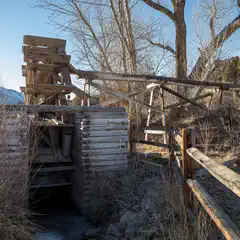 An old wooden water wheel stands beside a small stream, with a wooden trough attached to its edge.
