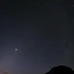 A shooting star streaks through a dark blue sky with many stars, a planet, and a mountain peak.