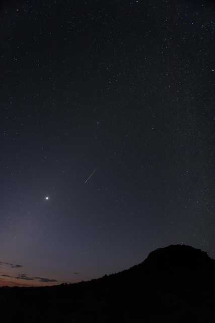 A shooting star streaks through a dark blue sky with many stars, a planet, and a mountain peak.