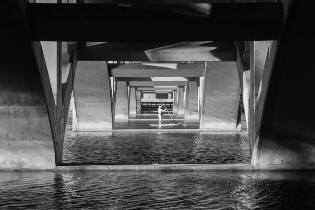 A person is paddleboarding under a concrete bridge with multiple supporting beams.