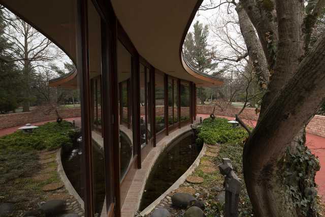 A modern house with a curved roof and large windows overlooking a garden with trees and a stream.