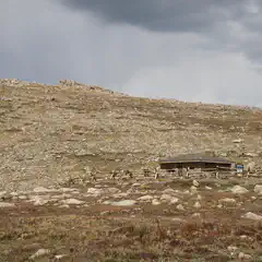 A small stone building stands in front of a rocky hillside under a cloudy sky.