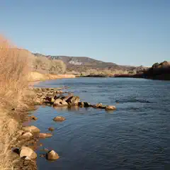 A tranquil river meandering through a landscape of dry grasses and shrubs with mountains in the distance against a clear blue sky.