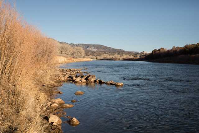 A tranquil river meandering through a landscape of dry grasses and shrubs with mountains in the distance against a clear blue sky.