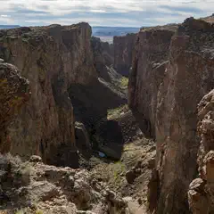 A rugged canyon landscape features towering rock formations and a narrow, winding gorge under a partly cloudy sky.
