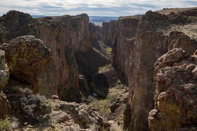 A rugged canyon landscape features towering rock formations and a narrow, winding gorge under a partly cloudy sky.