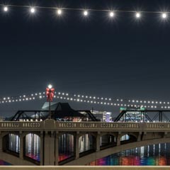 A bridge at night, illuminated by lights strung across its span and along its sides, with reflections of colorful lights on the water below.