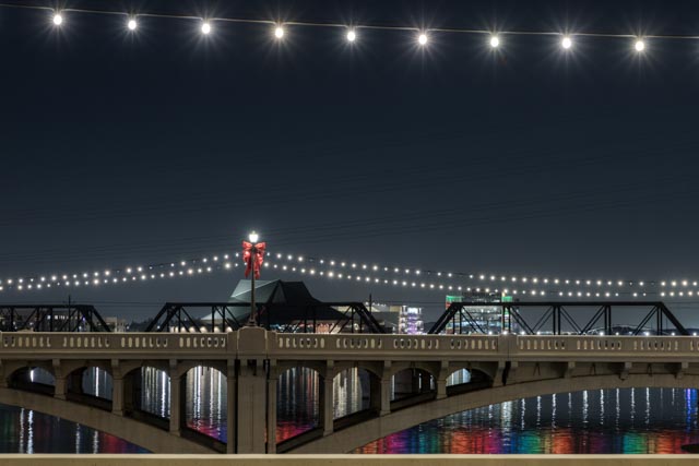 A bridge at night, illuminated by lights strung across its span and along its sides, with reflections of colorful lights on the water below.