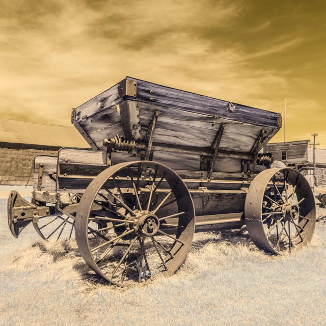 An old wooden wagon with large spoked wheels sits in a grassy field under a sepia-toned sky.