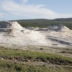 A cluster of geysers and hot springs is visible in a grassy area with trees in the background.